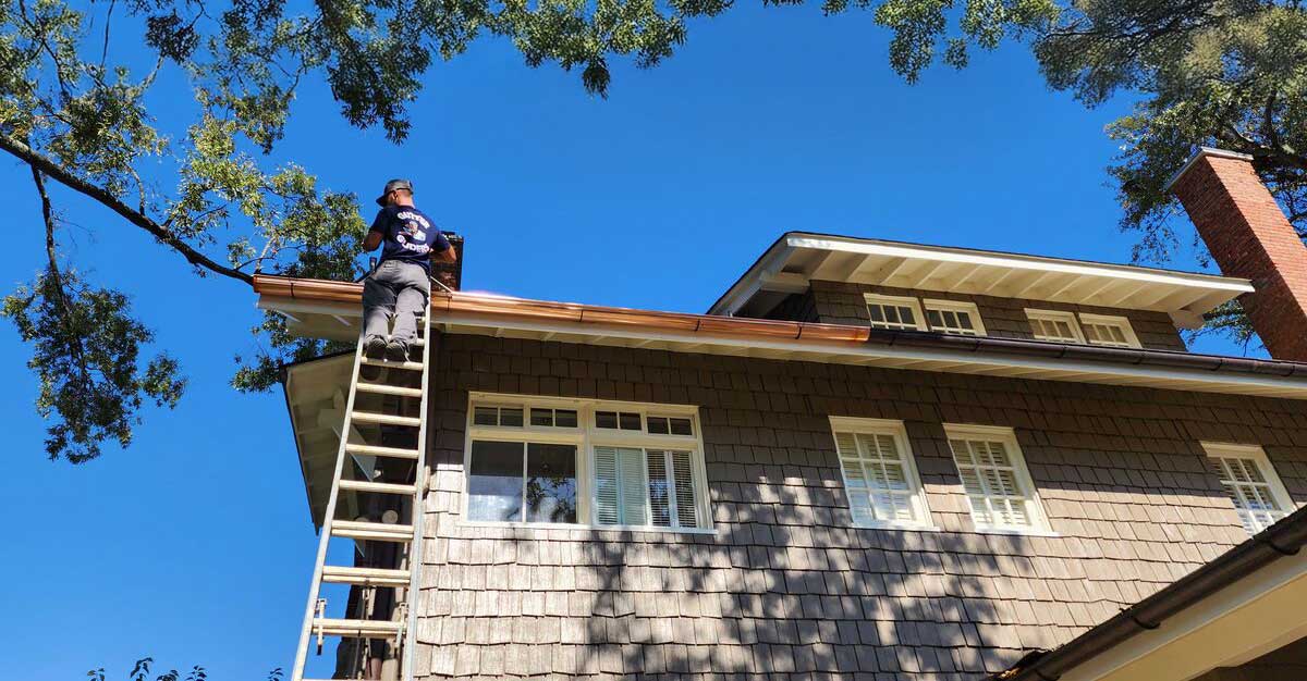 An upwards view of a home with a worker cleaning and repairing gutters from a ladder.